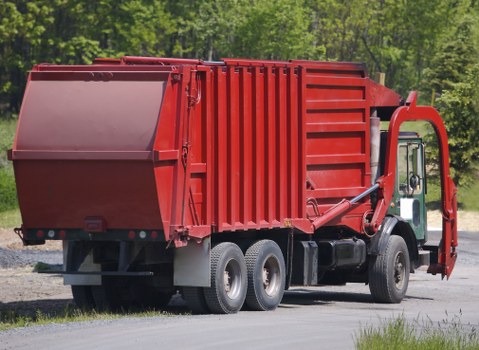 A large red skip loader truck parked on a paved driveway with a grassy verge and a backdrop of green trees, visible behind the vehicle. The truck features a prominent rectangular skip container with a smooth, painted metal surface and horizontal reinforcements, mounted on a black chassis with dual rear axles. The cab of the truck, positioned on the right, has a white roof and black side mirrors. The hydraulic arm and lifting mechanism are attached to the side of the skip, designed for transporting and unloading waste. The scene suggests a rural or suburban setting, with natural daylight illuminating the vehicle and surroundings. The presence of the skip truck indicates waste removal or rubbish collection services, typical of commercial waste management operations in areas like Norwood or nearby London boroughs, as handled by companies such as Commercial Waste Norwood.