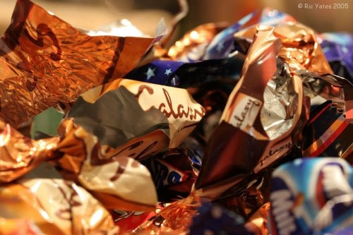 A close-up view of a pile of crumpled, glossy foil-wrapped chocolates and confectionery wrappers in various metallic shades of copper, silver, and gold, with some others in red, blue, and white colors. The wrappers are piled randomly, with some partially unfolded and overlapping each other, displaying blurred text and logos. The background is indistinct, suggesting an indoor setting possibly related to waste disposal or rubbish collection as might be seen in areas serviced by Commercial Waste Norwood for office and business rubbish removal. The image emphasizes the shiny, reflective surfaces of the wrappers and the chaotic arrangement typical of waste awaiting collection or disposal.