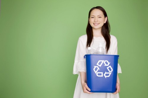 A young woman with long dark hair, wearing a white dress with lace details on the sleeves, is standing outdoors against a bright green background. She is smiling and holding a medium-sized blue recycling bin with a white recycling symbol printed on the front. The bin appears to be made of plastic, with a smooth surface and a slightly rounded top edge. The woman is positioned centrally, with her hands gripping the sides of the bin at waist level, and the bin is oriented upright. The environment suggests a clean, well-maintained area, possibly near a residential or commercial property in Norwood. This image visually supports waste management services, highlighting the focus on recycling and rubbish collection, particularly in the context of environmentally responsible office and business waste removal for local areas such as the inferred postcode and town served by Commercial Waste Norwood.