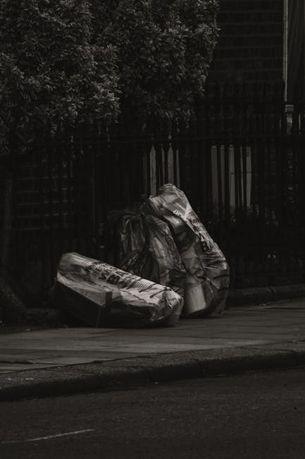 A collection of overflowing waste bins and scattered rubbish on a paved urban street, including a large grey mixed waste container filled with crumpled paper, cardboard, and plastic bags, with additio