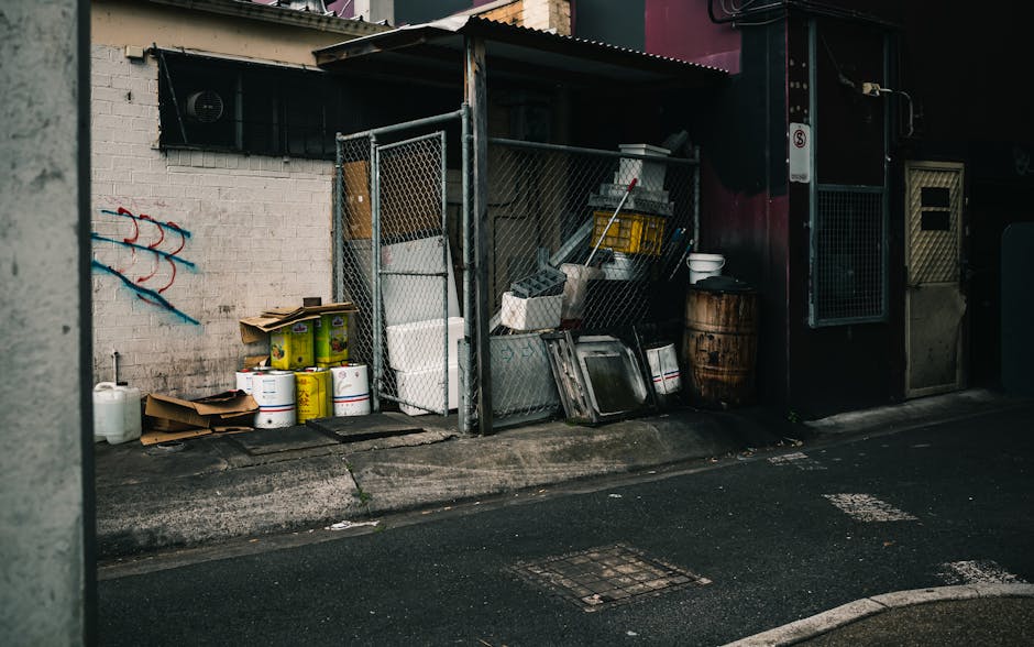 A designated outdoor rubbish collection area adjacent to a building, featuring a metal fence enclosure that contains various waste items. Inside the enclosure, there are large white plastic containers